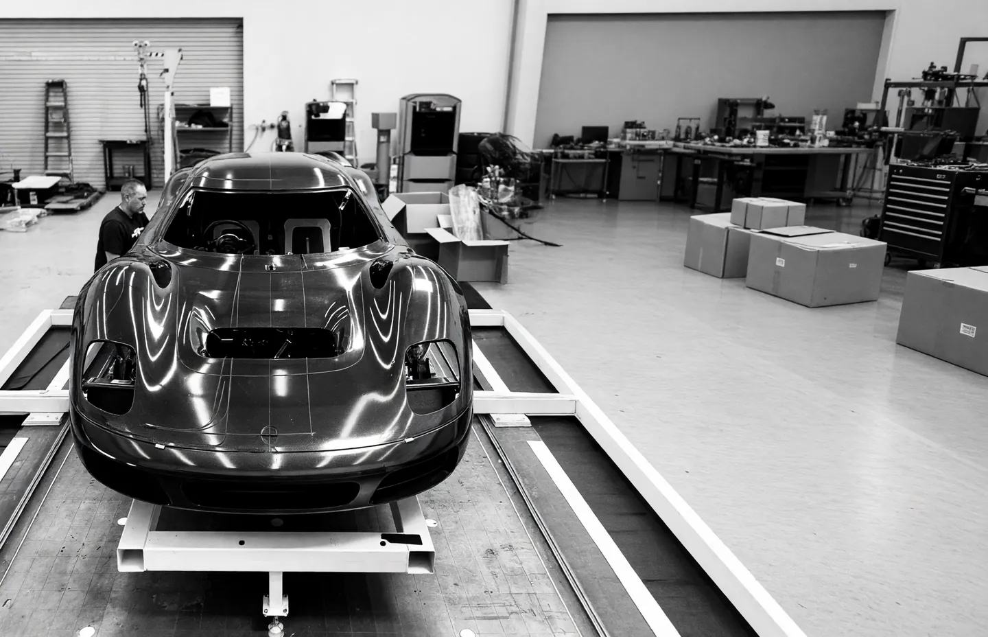 A carbon-fibre car body on a build frame in the workshop, seen from the front, with a technician working beside it.
